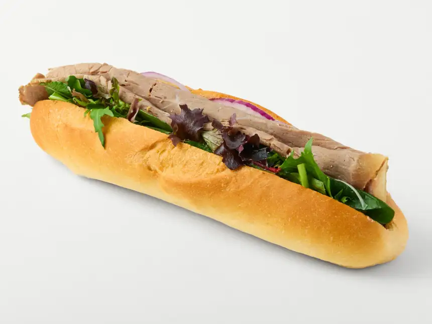 Filled baguette with salad leaves, sliced protein and red onion photographed against a clean white background