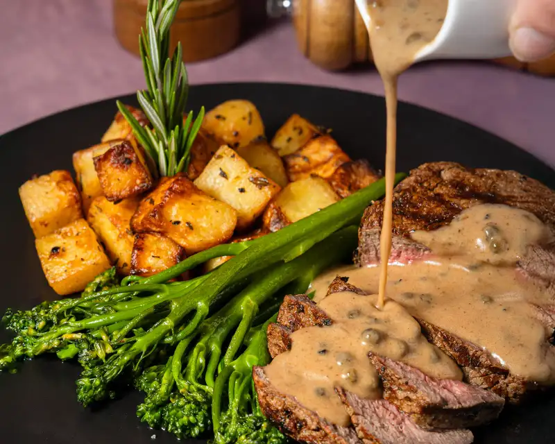 Action food photography of sauce being poured over a steak and broccolini dish, professionally styled.