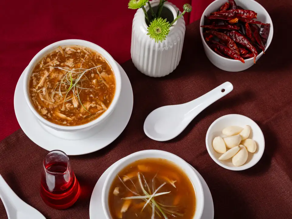 Hot and sour soup styled with dried chillies, garlic cloves, and a ceramic spoon on a red linen backdrop at Como Garden Restaurant