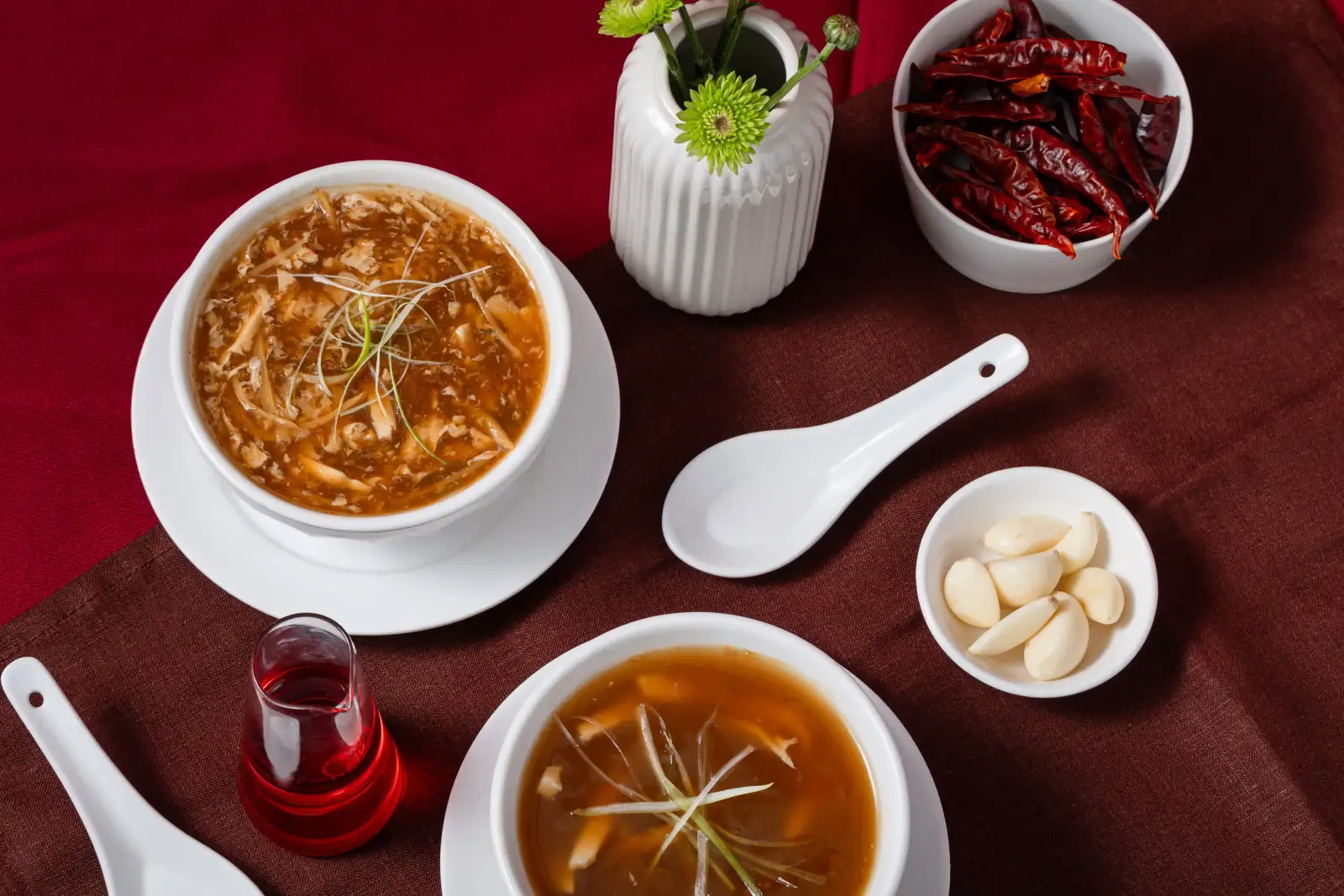 Hot and sour soup styled with dried chillies, garlic cloves, and a ceramic spoon on a red linen backdrop at Como Garden Restaurant