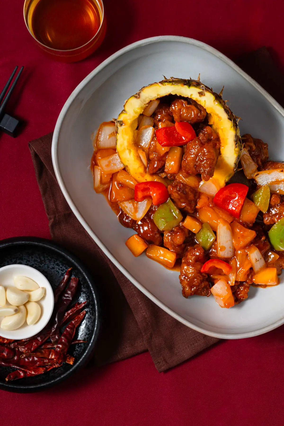 Overhead view of pineapple bowl stir-fry with chopsticks, tea, dried chillies, and garlic on a red backdrop at Como Garden Restaurant