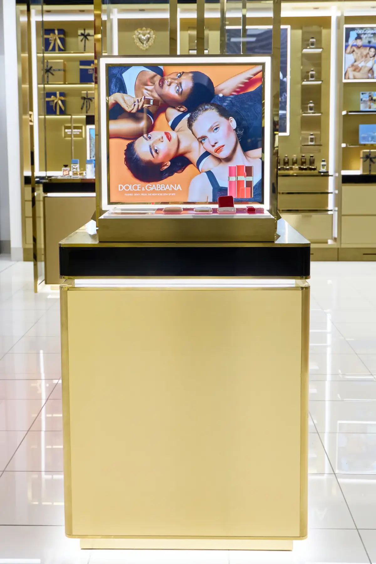Freestanding gold Dolce&Gabbana display plinth at Myer Perth with a backlit lip campaign panel and lipstick testers on the counter