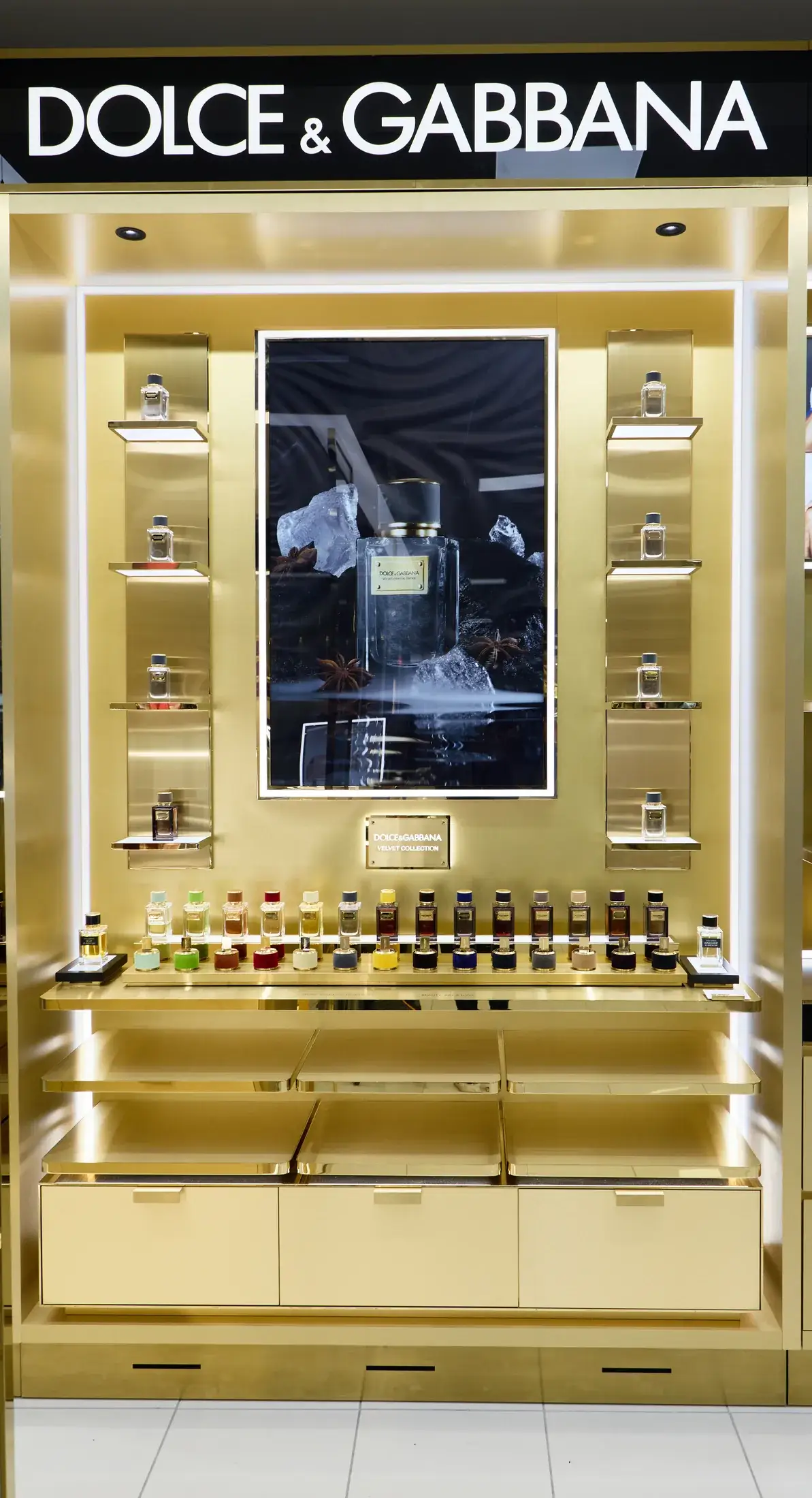 Dolce&Gabbana Velvet Collection fragrance wall at Myer Perth with backlit gold shelving and a full row of coloured perfume bottles