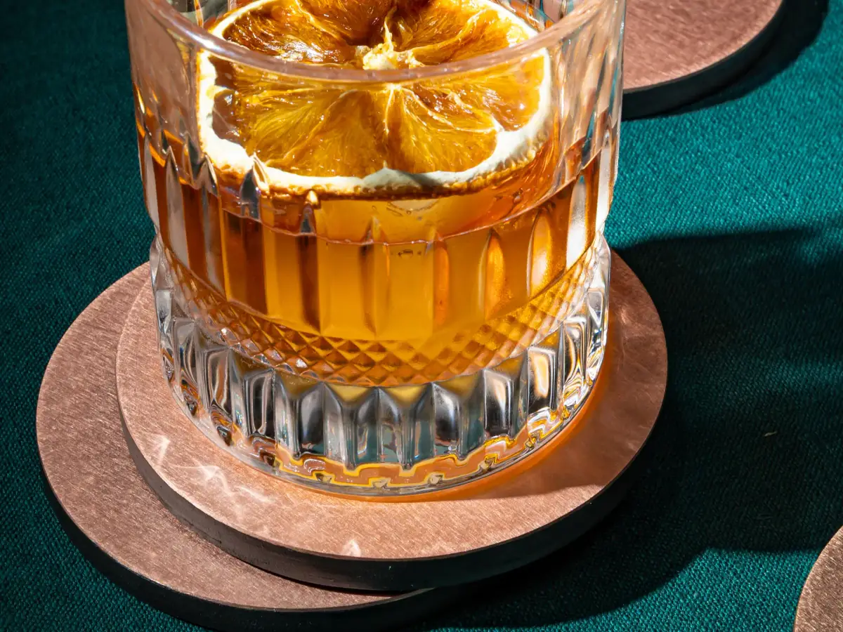 Whisky glass with dried orange slice resting on a brushed copper coaster close-up