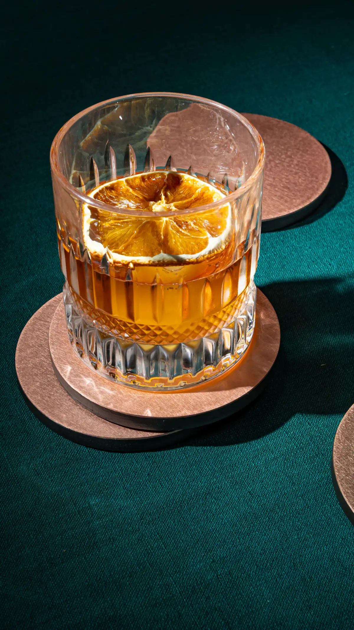 Whisky glass with dried orange slice resting on a brushed copper coaster close-up