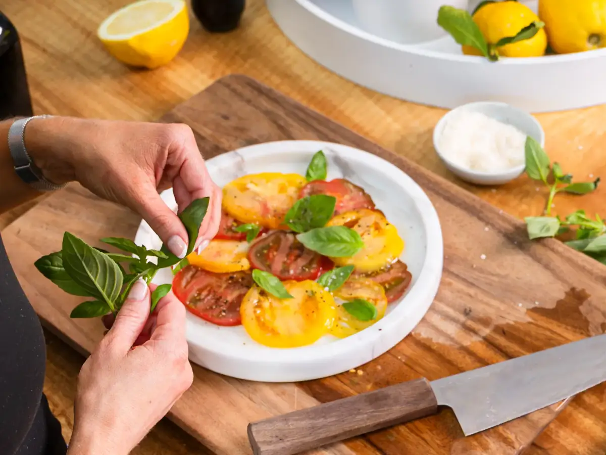 Hands garnishing an heirloom tomato salad on a wooden board with fresh basil and lemons nearby