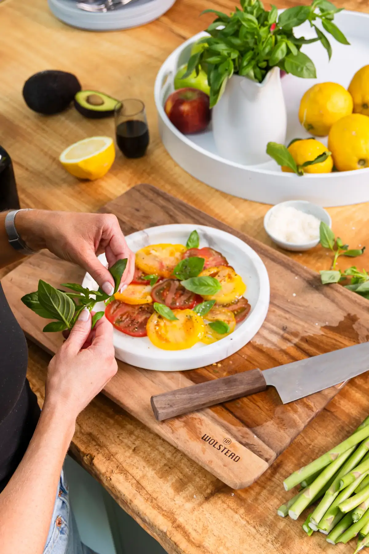 Hands garnishing an heirloom tomato salad on a wooden board with fresh basil and lemons nearby