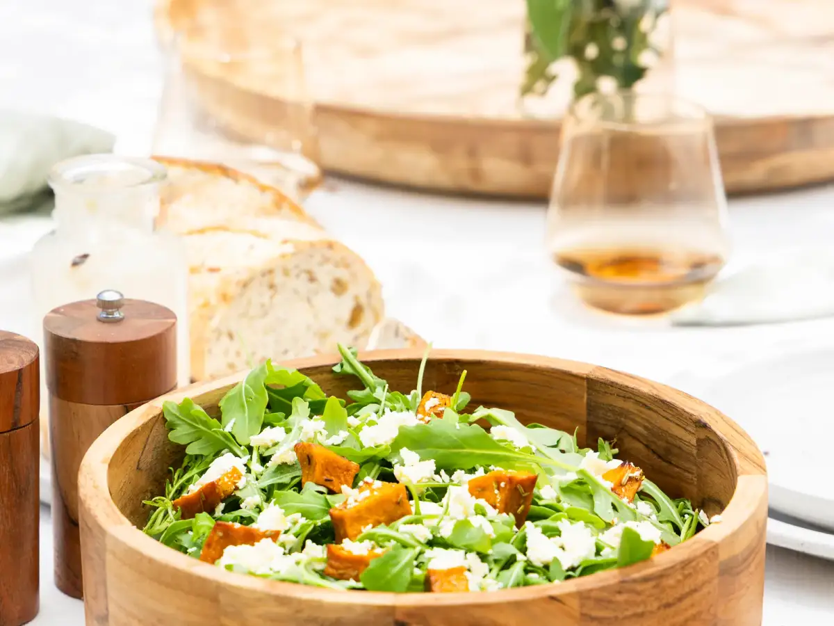 Wooden salad bowl filled with leafy greens, feta, and roast pumpkin on a bright styled table