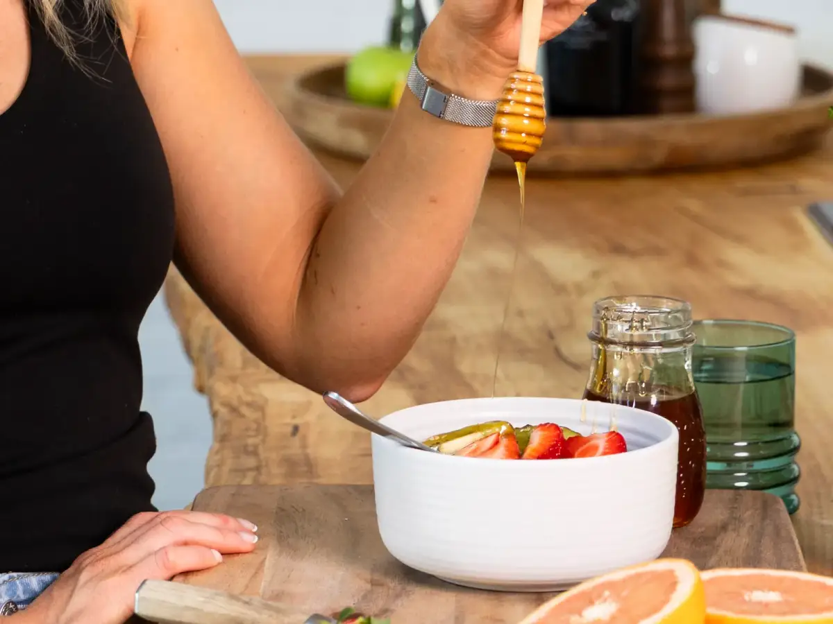 Honey drizzling into a yoghurt bowl topped with strawberries, with grapefruit and kiwi on the bench