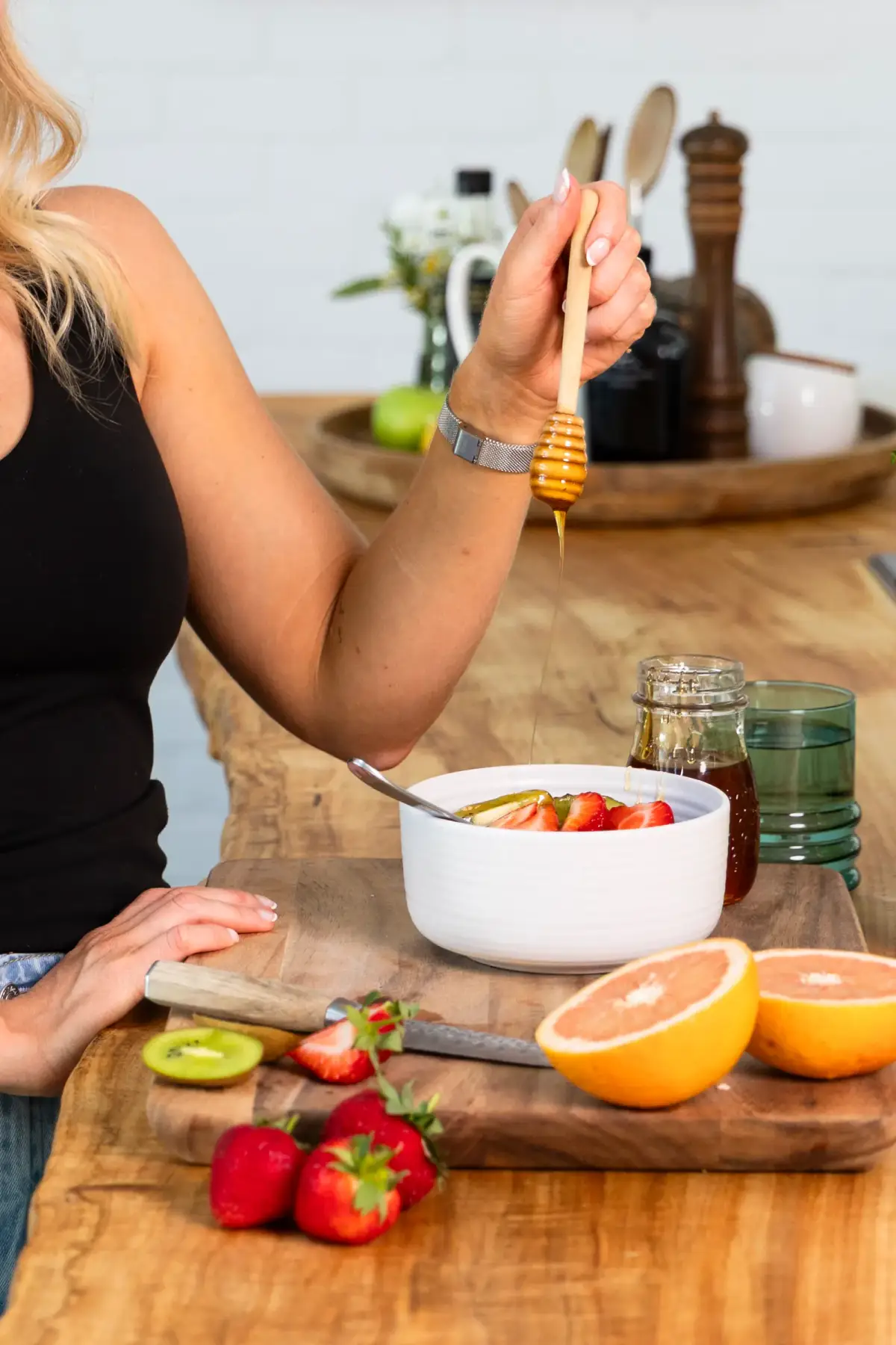 Honey drizzling into a yoghurt bowl topped with strawberries, with grapefruit and kiwi on the bench
