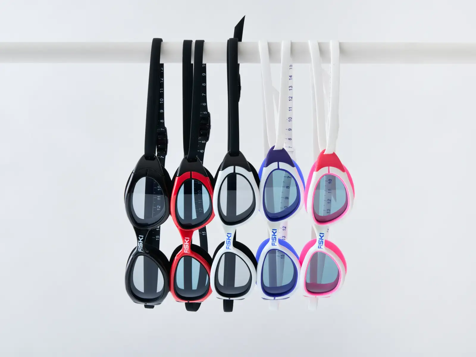Five pairs of Fiski swimming goggles in black, red, white, blue and pink hanging from a white rail against a clean white background
