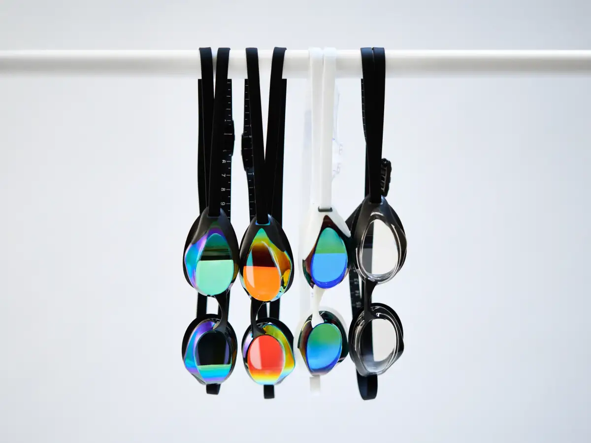 Range of Fiski swimming goggles with iridescent mirrored lenses in green, orange, blue and silver hanging from a white rail