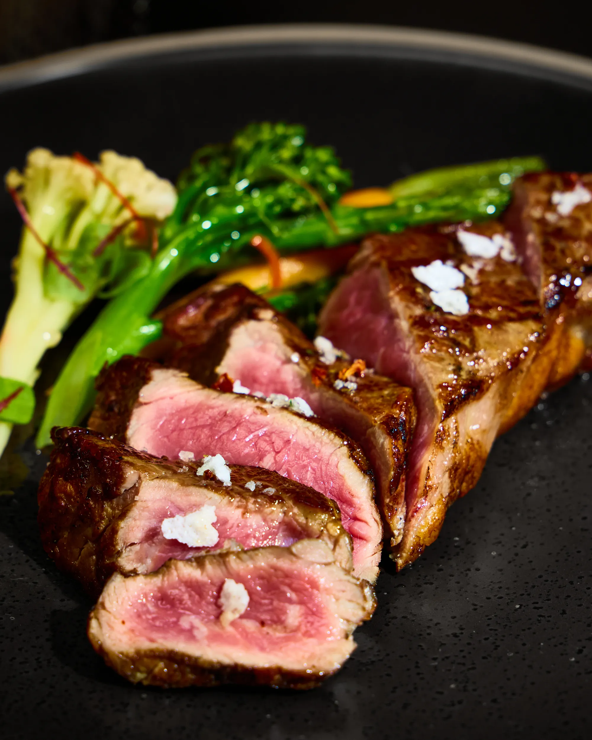 Sliced grilled steak with broccolini and sauce on a black plate
