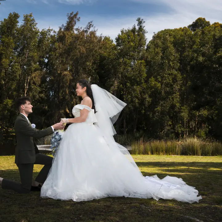 Playful wedding portrait of the groom kneeling and holding the bride's hands beside the lake.