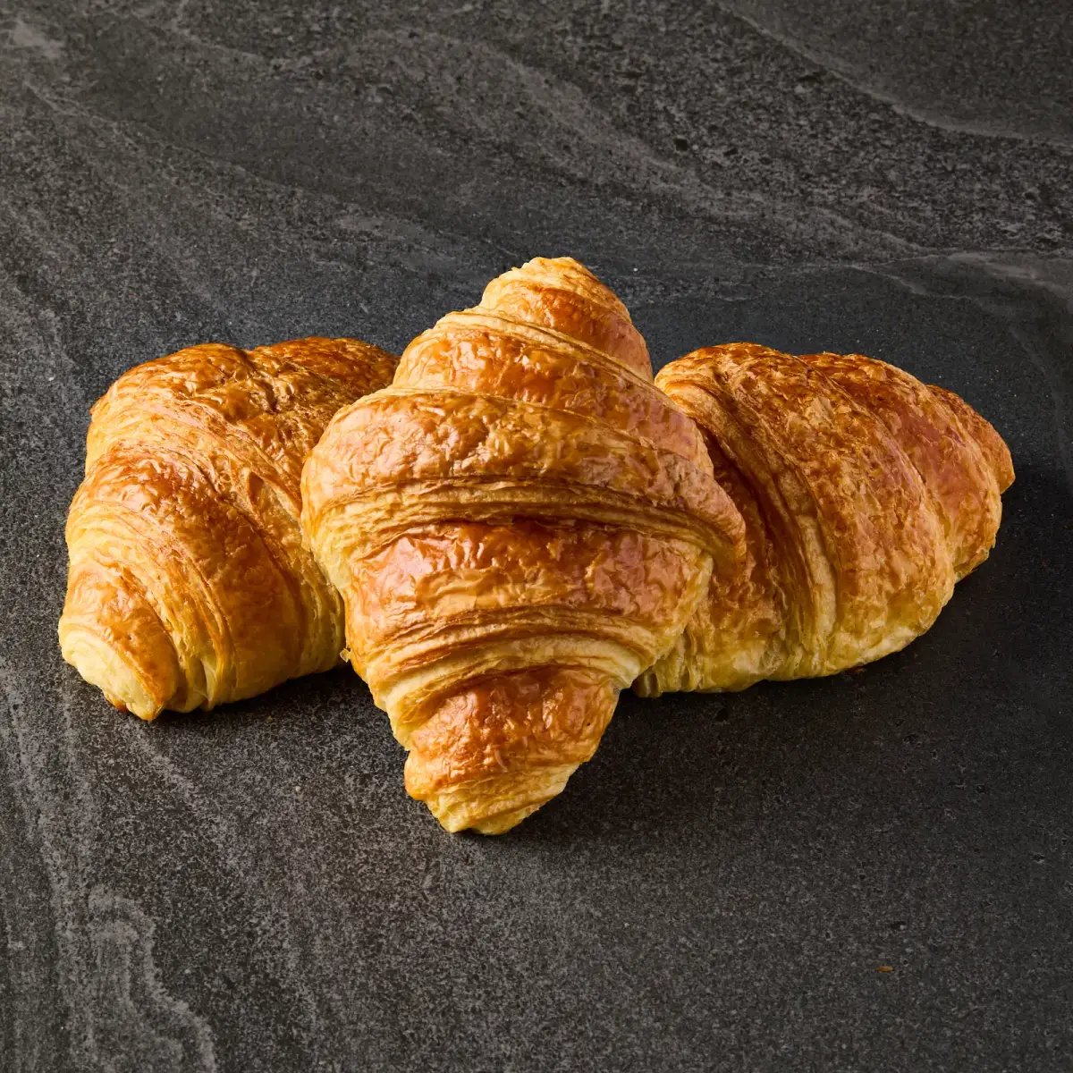 Three golden, flaky Il Granino butter croissants arranged on a dark stone surface for bakery product photography