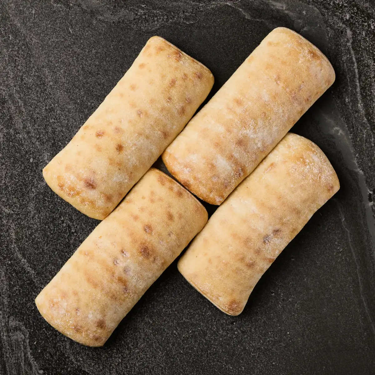 Four floured Il Granino ciabatta rolls photographed from above on a dark stone surface