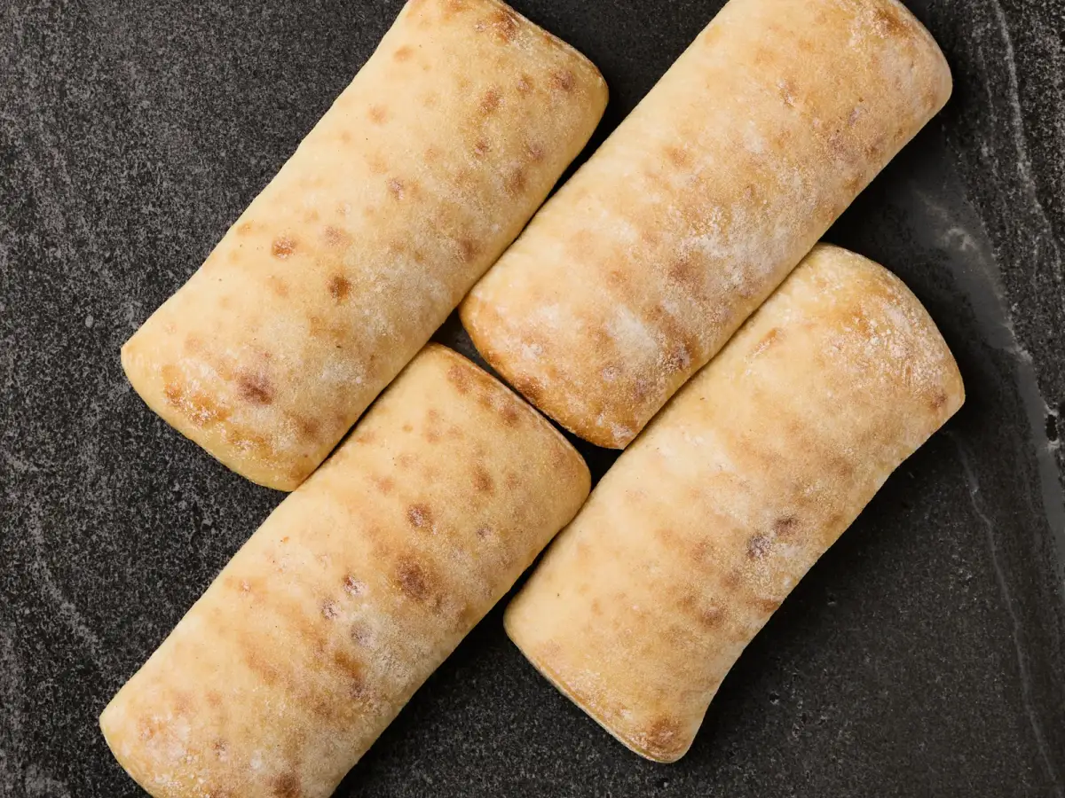 Four floured Il Granino ciabatta rolls photographed from above on a dark stone surface
