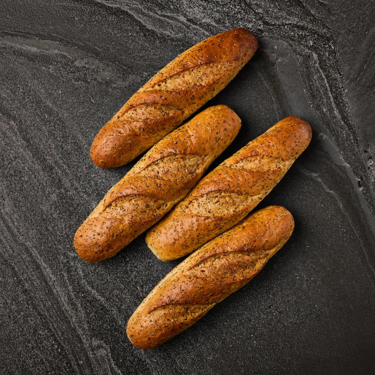 Four Il Granino seeded baguettes with poppy and sesame crust arranged overhead on a dark stone surface