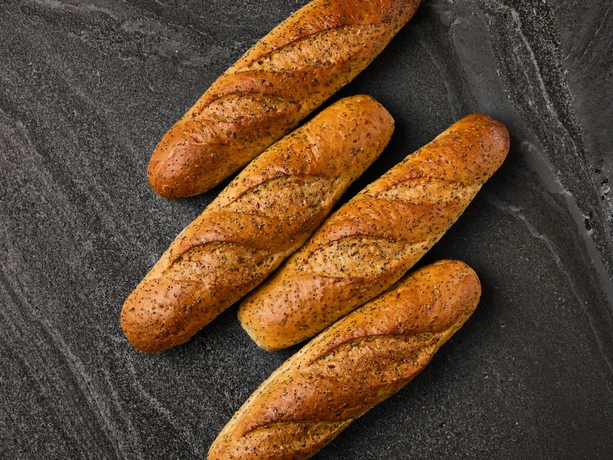 Four Il Granino seeded baguettes with poppy and sesame crust arranged overhead on a dark stone surface