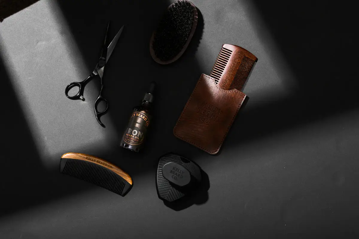 Kelly Beard Co beard oil, wooden combs, brush, scissors, and grooming accessories arranged on a dark studio flatlay