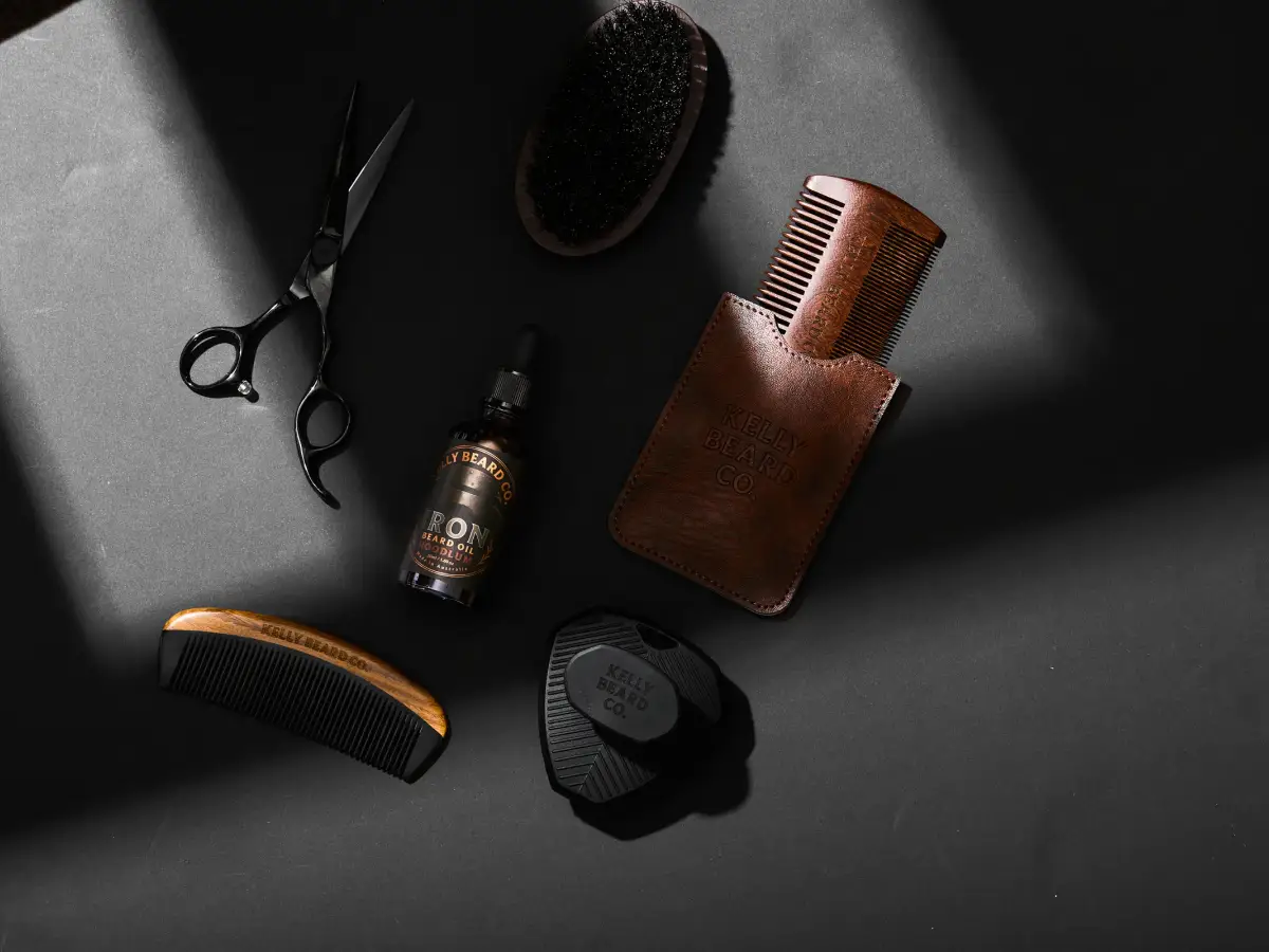 Kelly Beard Co beard oil, wooden combs, brush, scissors, and grooming accessories arranged on a dark studio flatlay