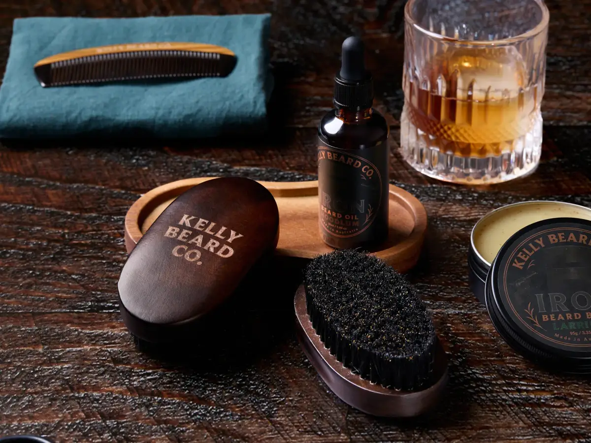 Kelly Beard Co beard oil, beard balm, wooden comb, brush, and whisky glasses styled on a dark timber surface