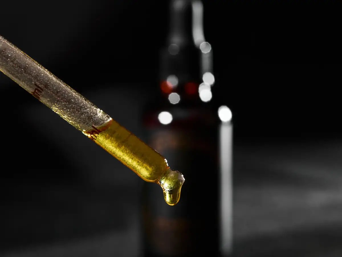 Close-up of golden Kelly Beard Co Hoodlum beard oil dripping from a glass dropper with product bottle blurred in the background