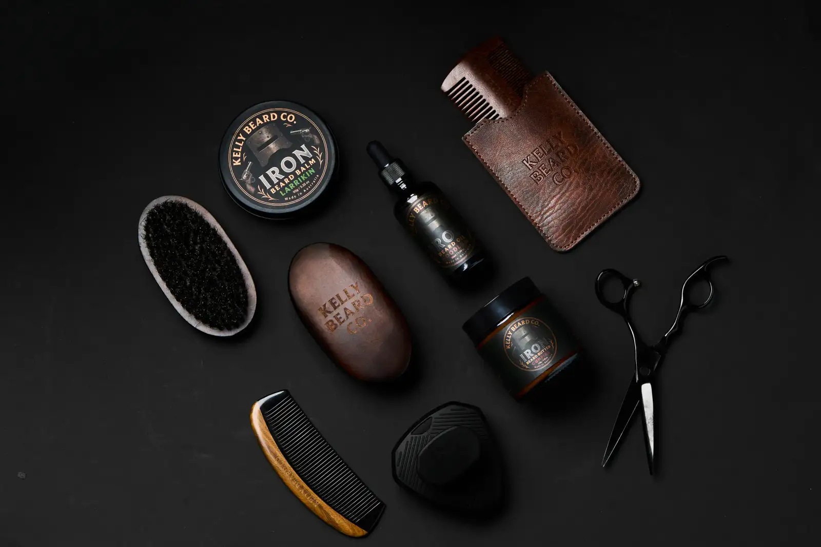 Kelly Beard Co Iron Beard balm, beard oil, combs, brushes, and grooming tools arranged on a dark flatlay for premium product photography