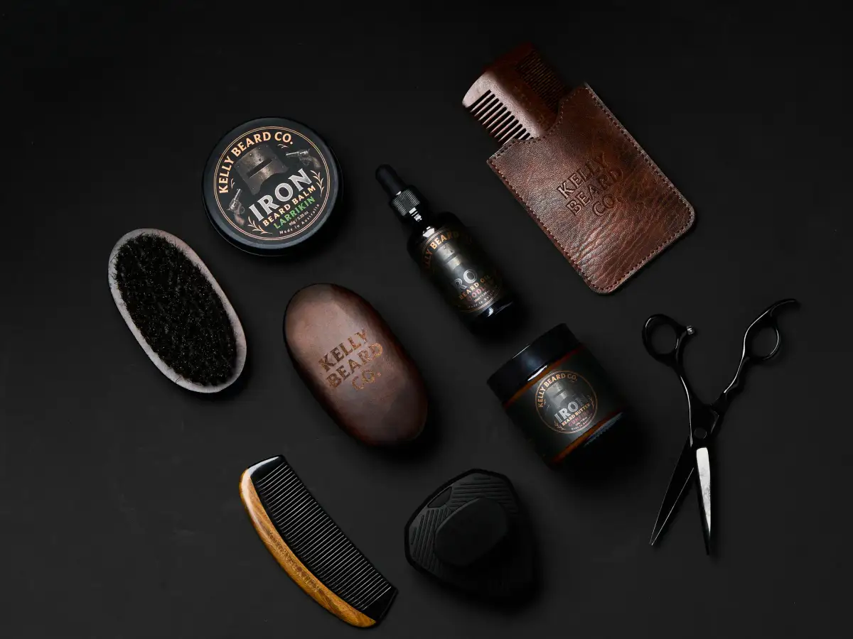 Kelly Beard Co Iron Beard balm, beard oil, combs, brushes, and grooming tools arranged on a dark flatlay for premium product photography