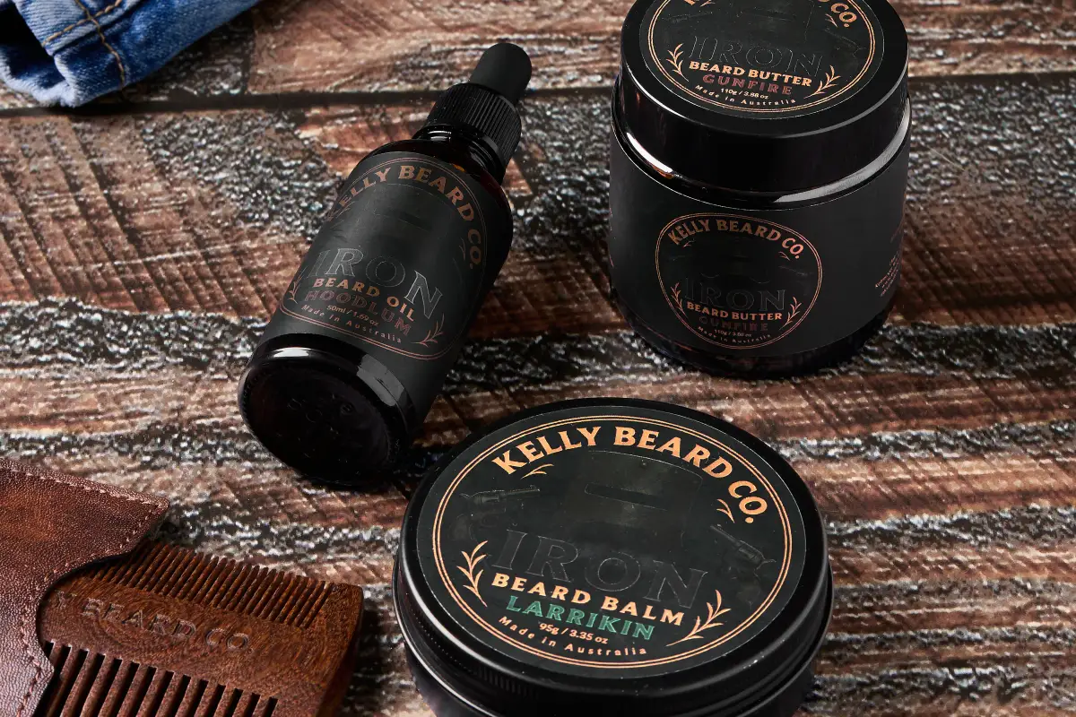 Kelly Beard Co Iron Beard oil, beard butter, and beard balm with a wooden comb styled on a rustic dark timber surface with denim