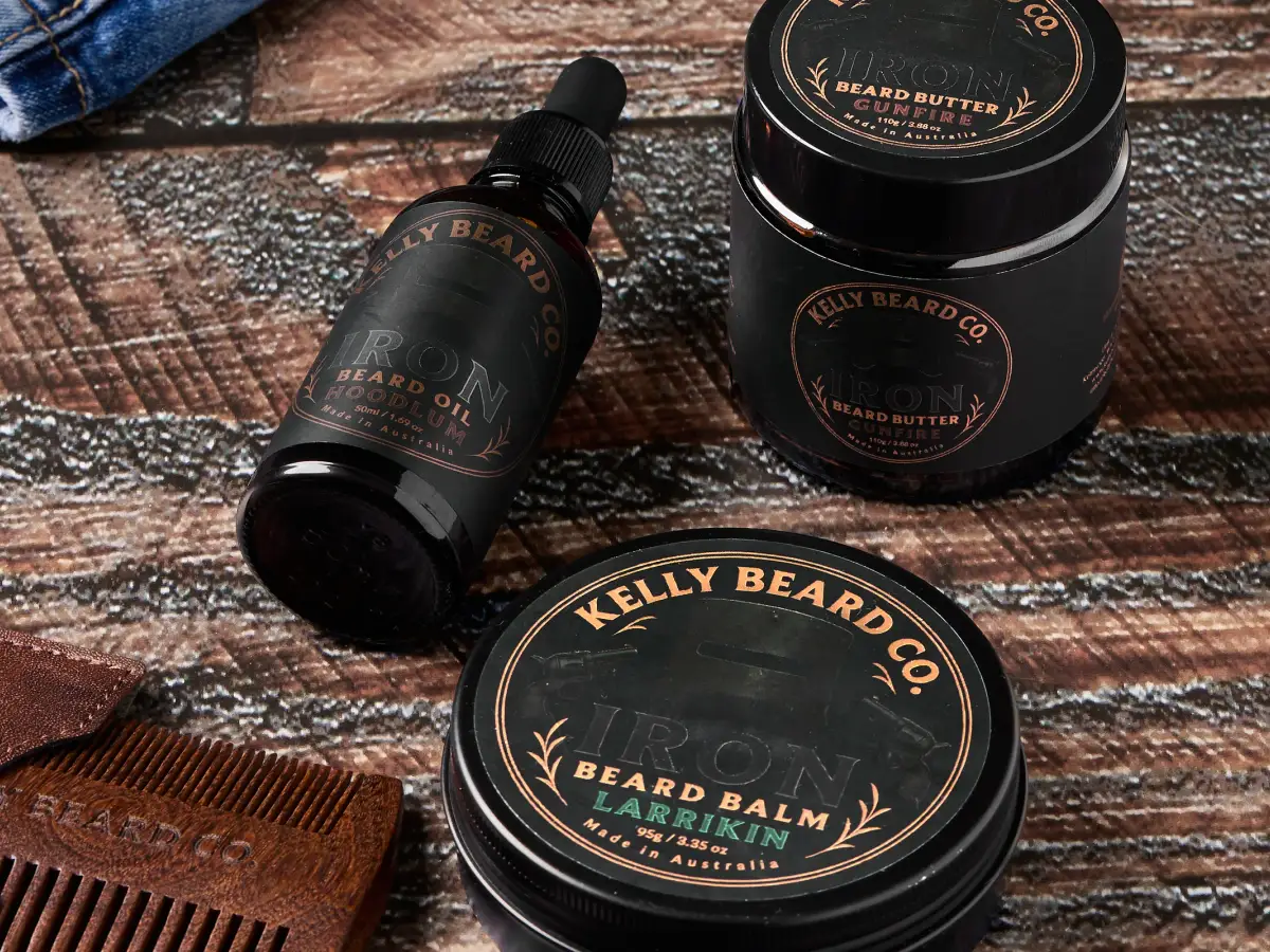 Kelly Beard Co Iron Beard oil, beard butter, and beard balm with a wooden comb styled on a rustic dark timber surface with denim