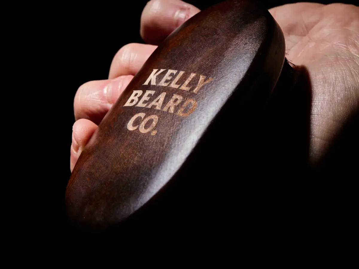 Close-up of a Kelly Beard Co wooden beard brush held in hand showing engraved branding and polished timber finish