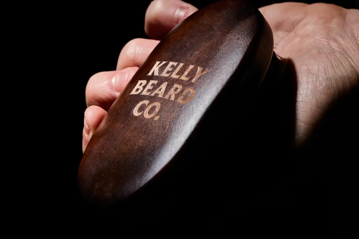 Close-up of a Kelly Beard Co wooden beard brush held in hand showing engraved branding and polished timber finish