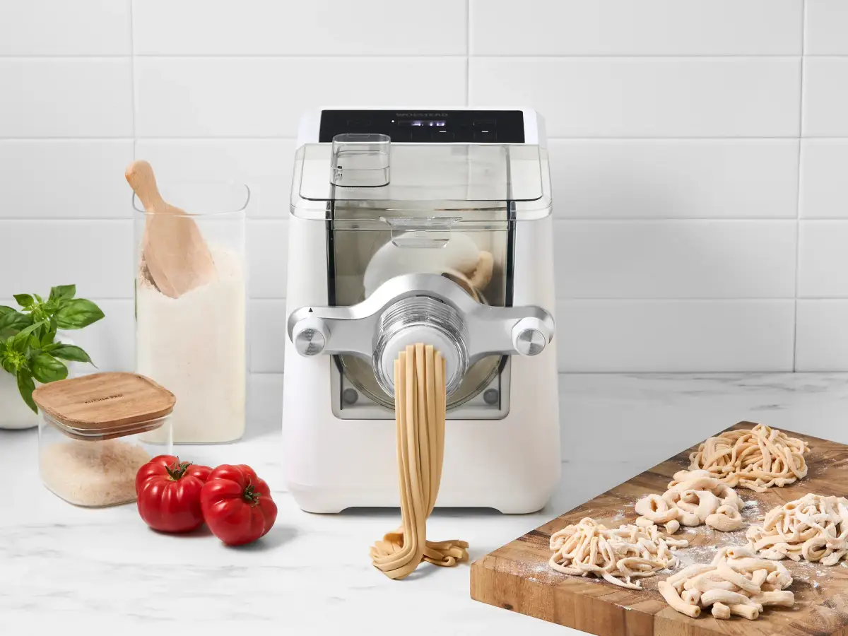White Wolstead Forma pasta maker extruding fresh pasta beside flour, tomatoes, basil, and assorted pasta shapes