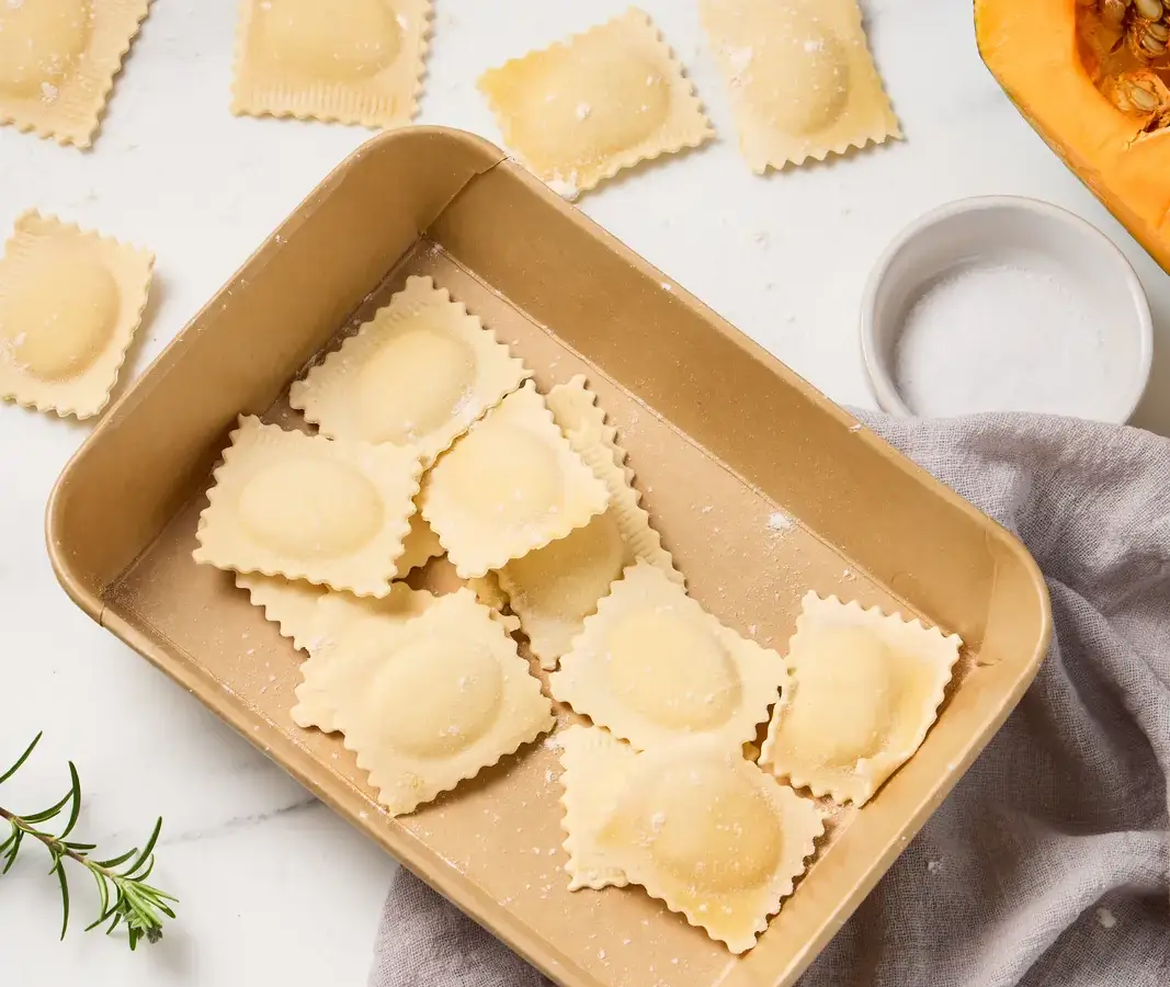 Fresh La Delizia Lab pumpkin ravioli arranged in a kraft tray with sage, walnuts, and a roasted pumpkin half on marble