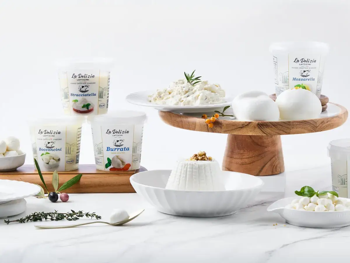 Styled spread of La Delizia Latticini handcrafted cheese with branded burrata, stracciatella, bocconcini, and mozzarella tubs, fresh mozzarella on a wooden stand, and ricotta on marble