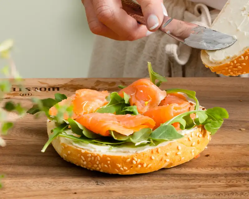 Bright lifestyle photography showing hands spreading cream cheese on a smoked salmon bagel.