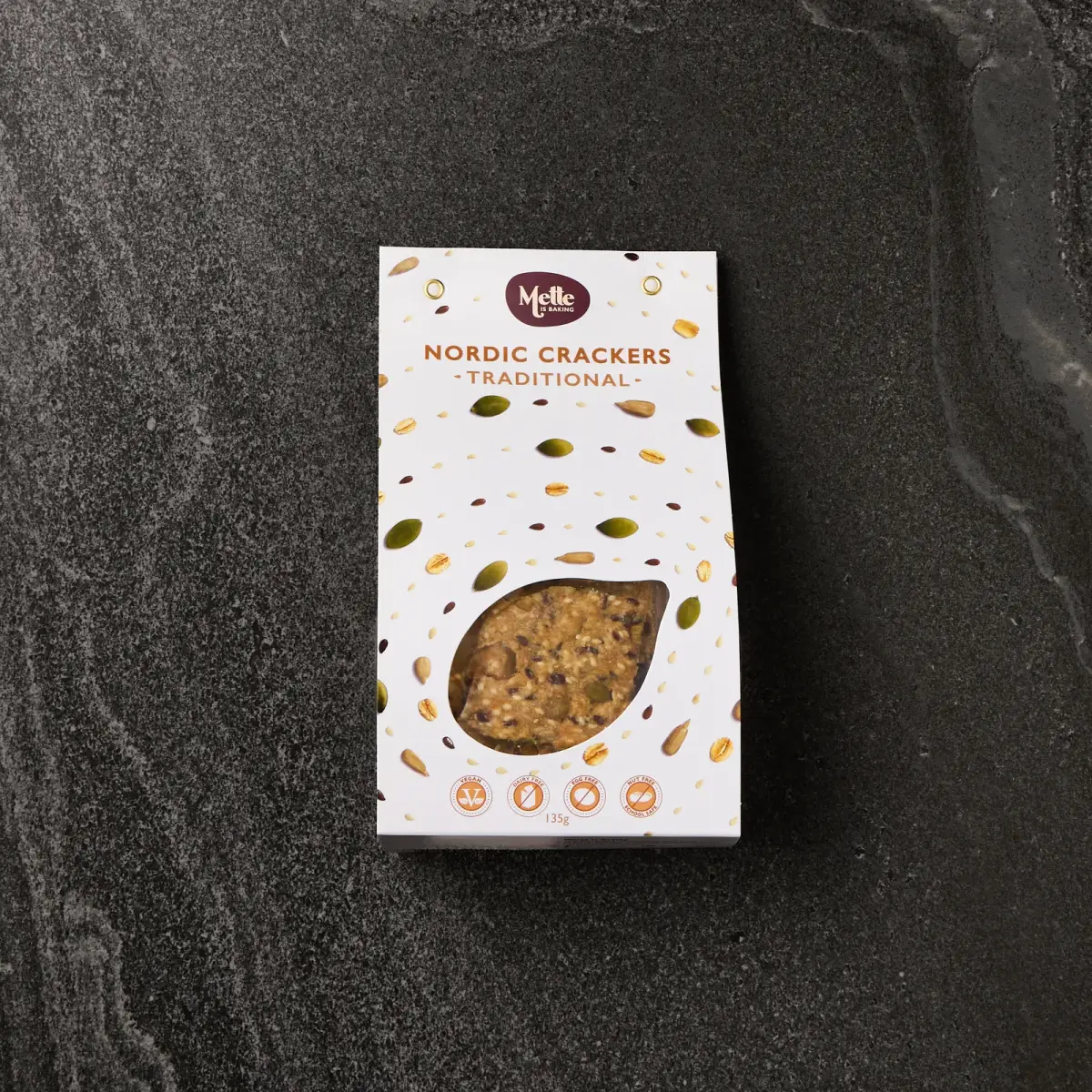 Mette is Baking Traditional Nordic Crackers retail box with seed-print packaging photographed on a dark stone surface