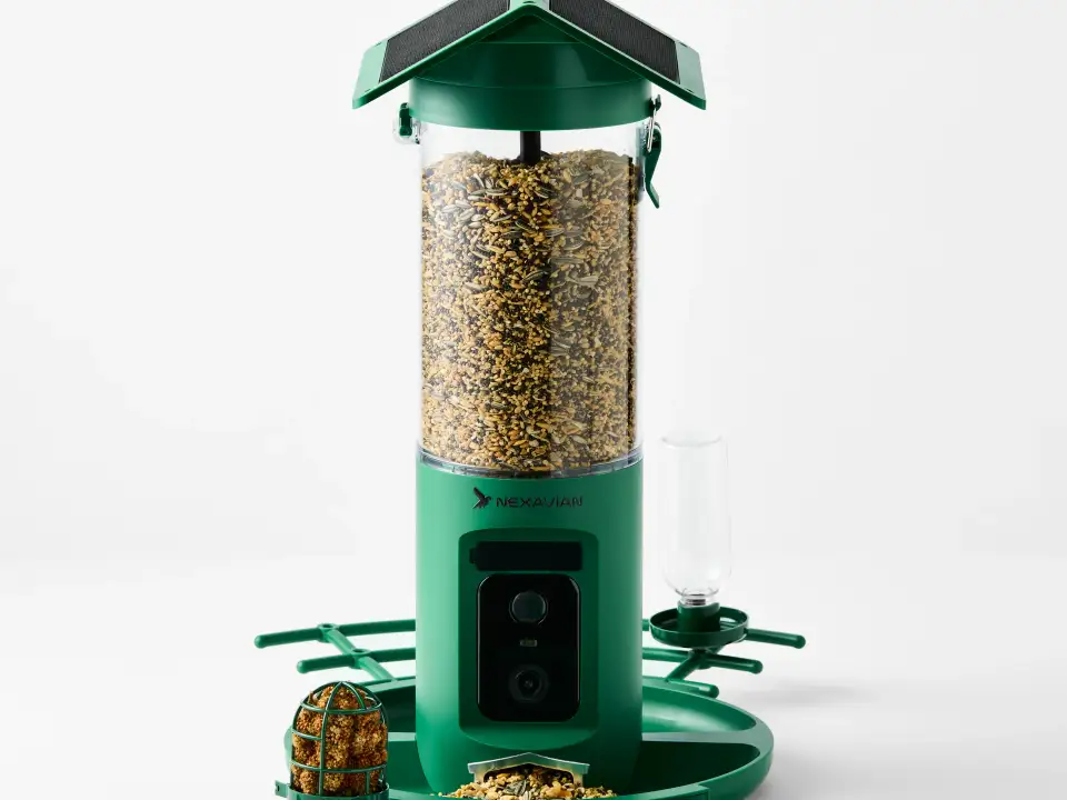 Green Nexavian camera bird feeder filled with seed, with its roof, front camera and feeding attachments visible on a light background