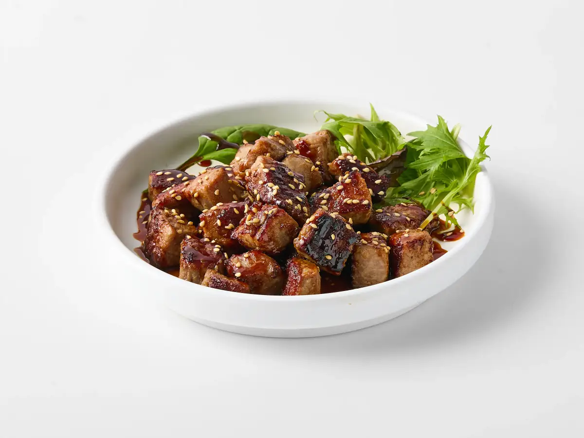 Omasa Japanese Restaurant beef dish glazed with dark sauce and sesame seeds, plated with mixed greens on a white background