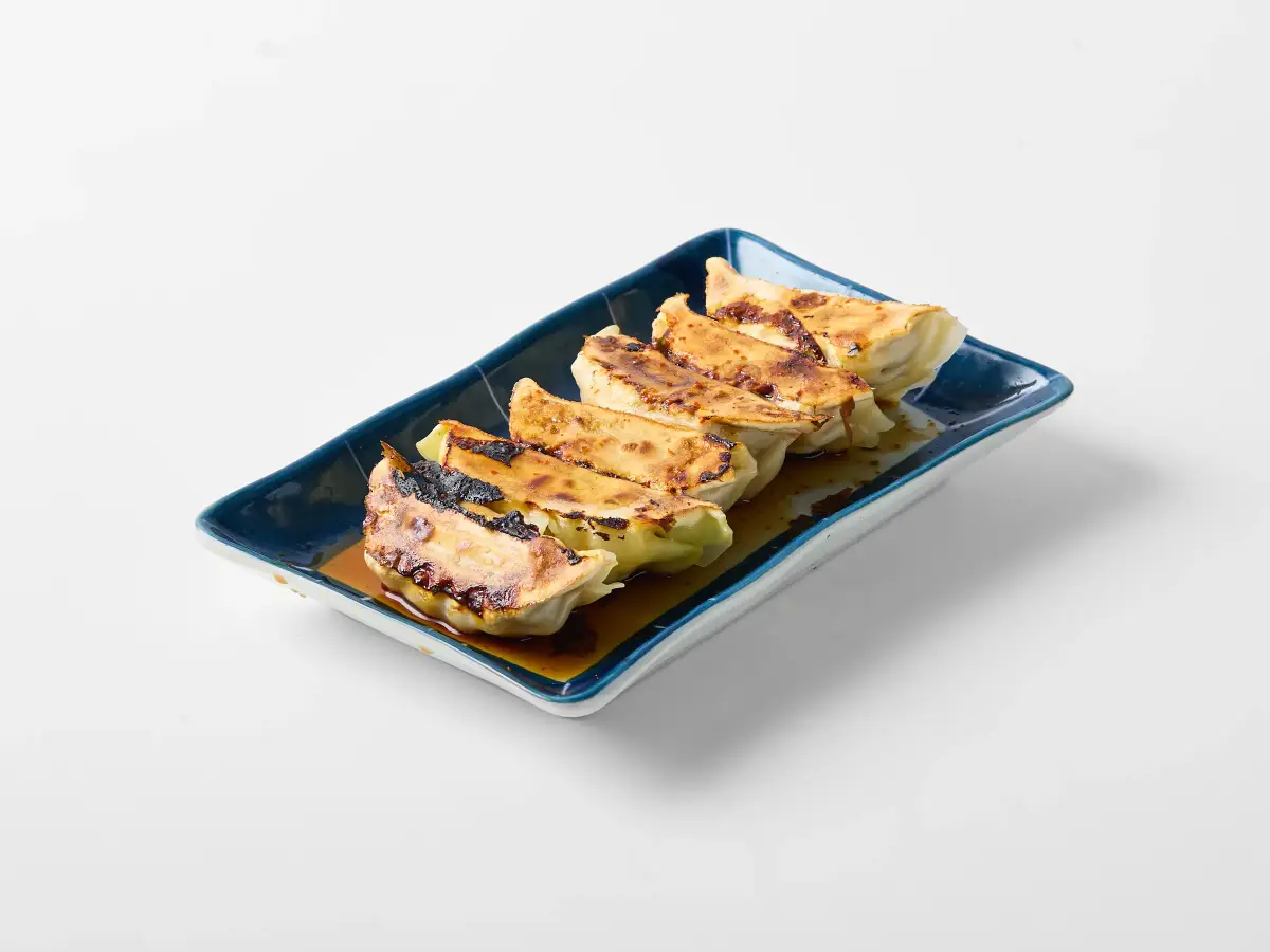Pan-fried gyoza from Omasa Japanese Restaurant lined on a rectangular plate with dipping sauce on a white background