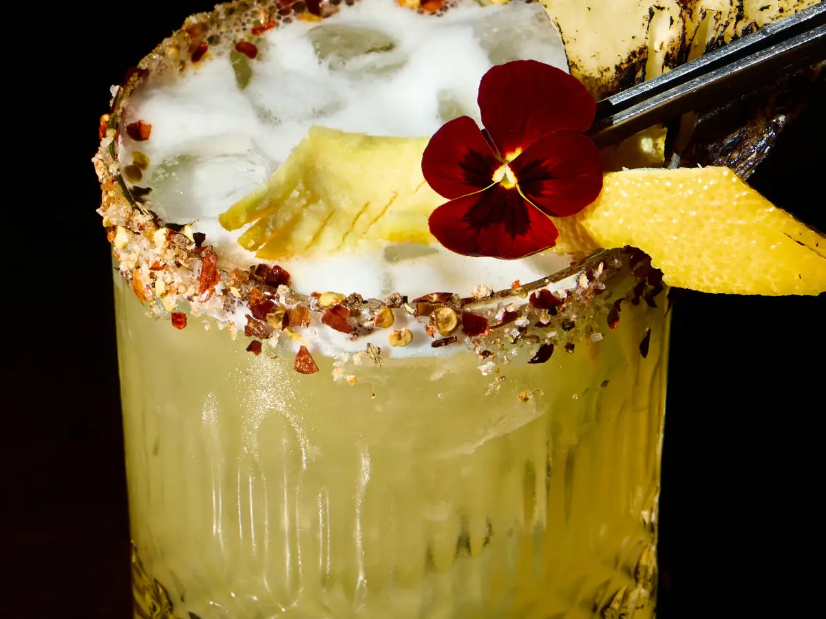 Tropical margarita-style cocktail at T.O.T.O Bar with a spiced rim, pineapple garnish, flower, and dark background