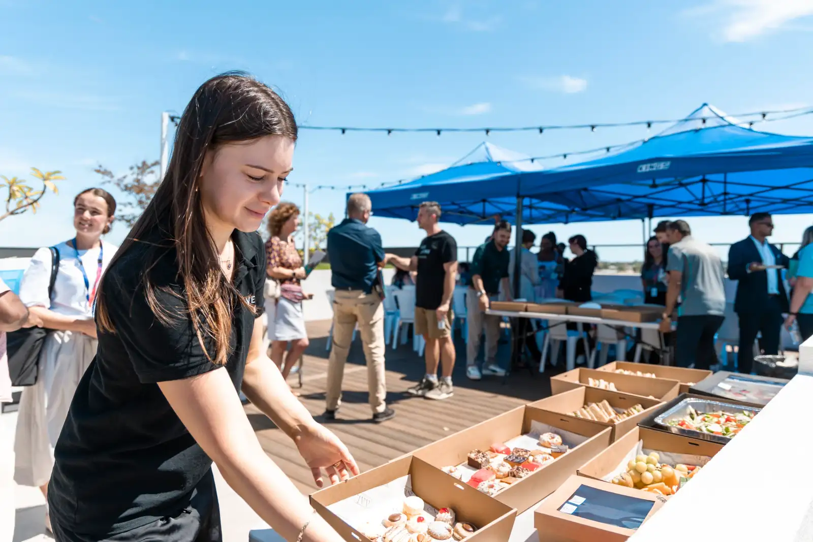 Vera Living staff member arranging catering boxes on a rooftop table while guests gather under blue marquees in Cannington
