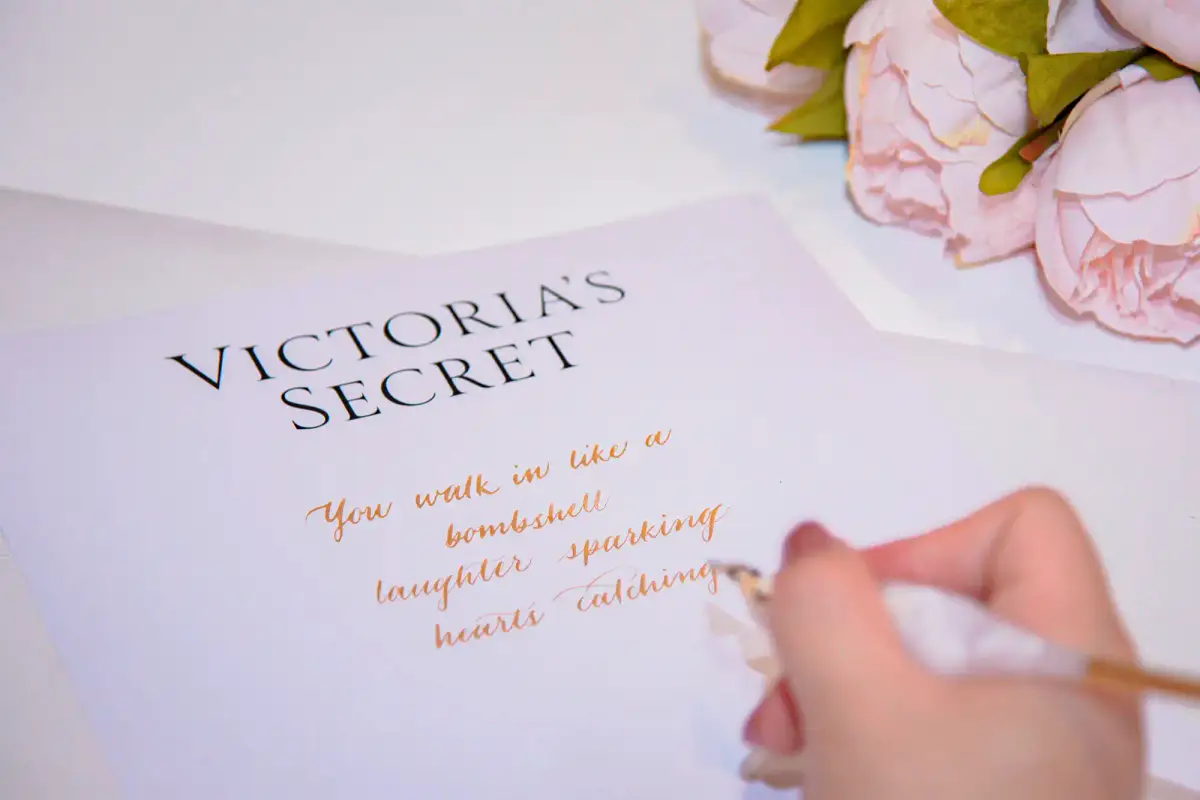 Hand writing gold calligraphy on Victoria's Secret branded stationery beside pink peonies