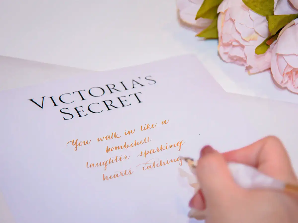 Hand writing gold calligraphy on Victoria's Secret branded stationery beside pink peonies