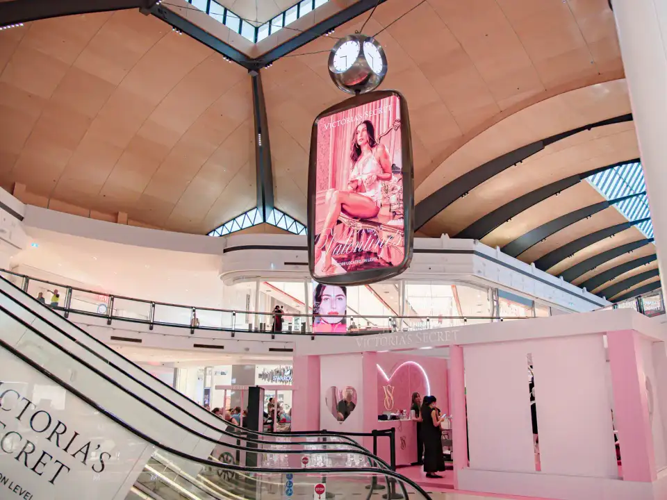 Wide atrium view of Karrinyup Shopping Centre showing the Victoria's Secret store opening with digital signage, escalators, and pink branded pop-up