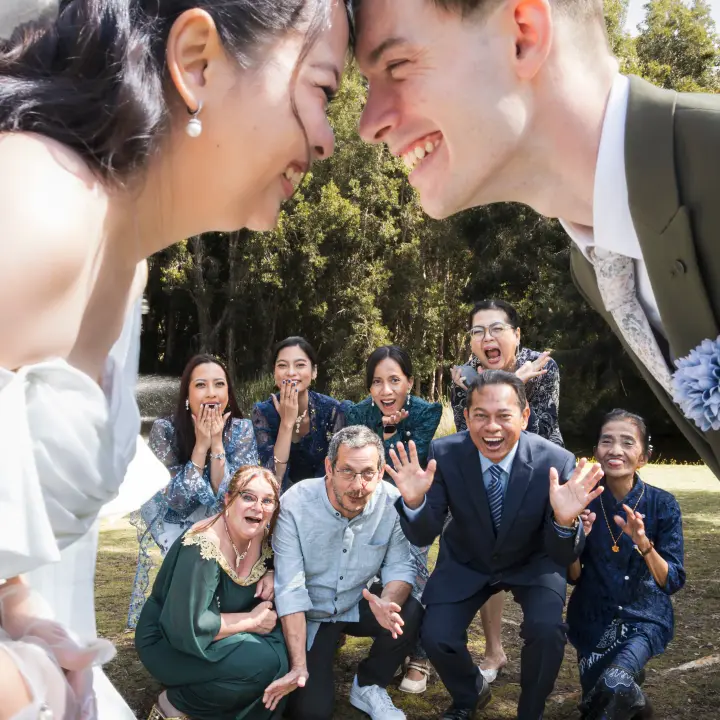 Bride and groom smiling forehead to forehead while family react behind them during an outdoor wedding portrait.