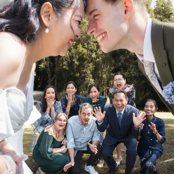 Newlyweds sharing a close portrait while family react behind them.
