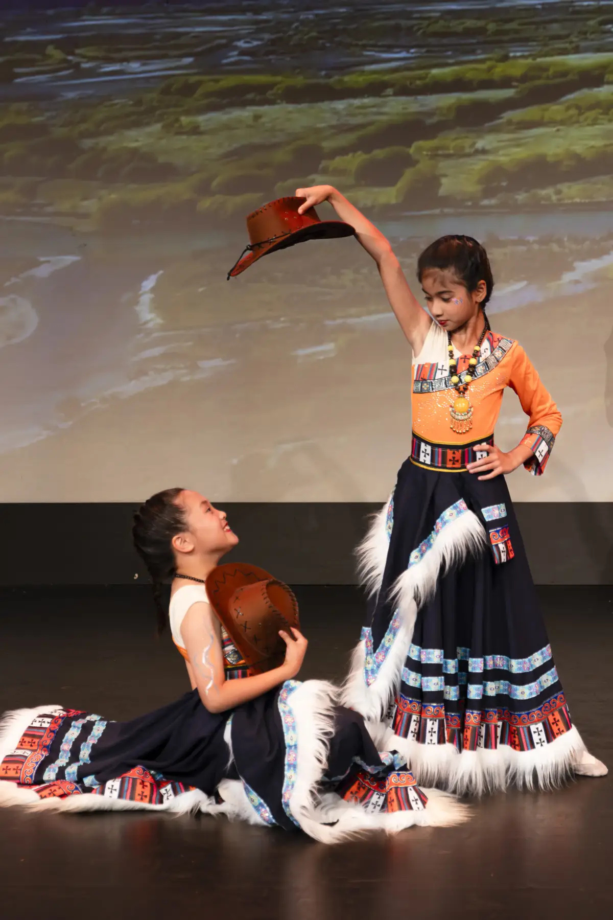 Two young dancers in patterned costumes posing in a duet performance with one holding a hat overhead