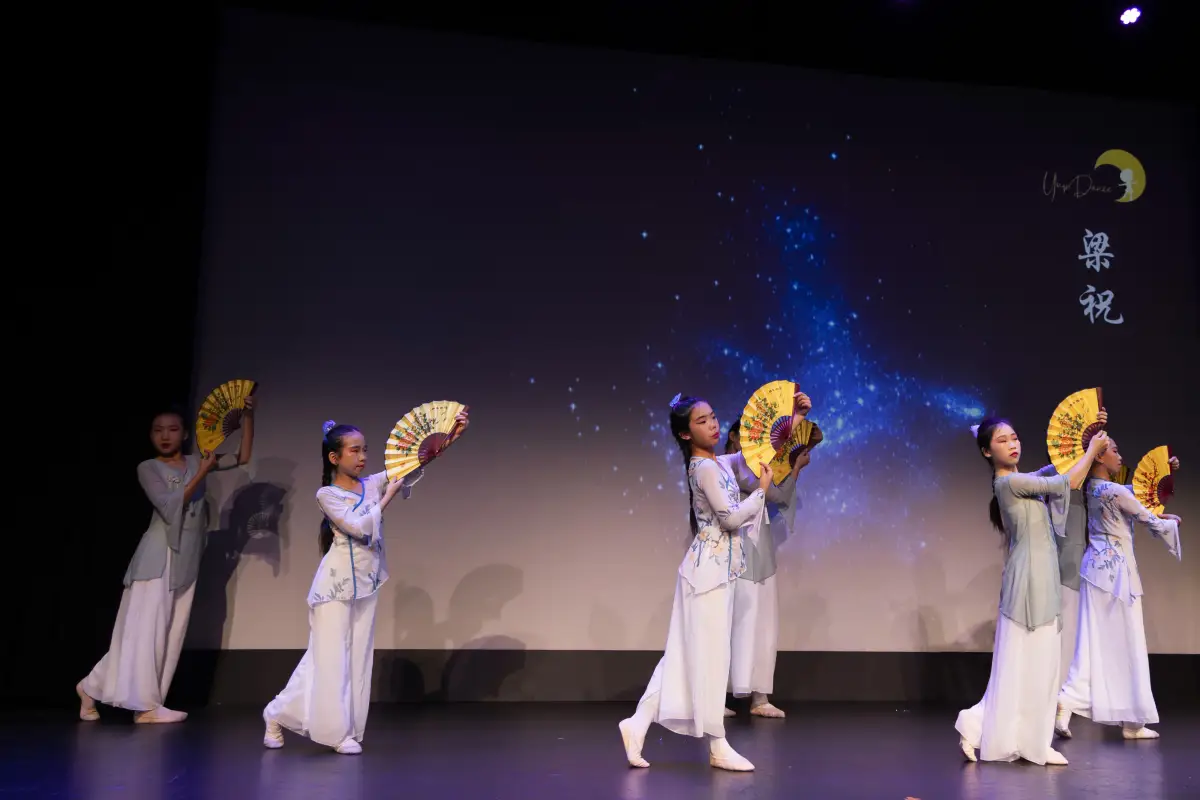 Five students in pale blue costumes performing a fan routine under a starry stage backdrop